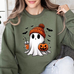 Op de afbeelding: Olijfgroene sweatshirt met een cartoon spookontwerp. Het spook draagt een oranje muts en houdt een pompoen en een drankje vast. Zwarte vleermuizen en sterren zijn verspreid rond het spook. Het spook heeft grote zwarte ogen en een vriendelijke uitdrukking.