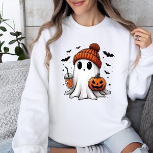 Op de afbeelding: Wit sweatshirt met een cartoongeest met een oranje gebreide muts. De geest houdt een pompoen en een drankje met een rietje vast. Zwarte vleermuizen en sterren omringen de geest en cre&euml;ren een Halloween-thema.