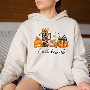 May include: Cream-colored hoodie with a graphic of fall-themed items: an orange beanie, a leopard-print tumbler, brown boots, and a pumpkin with a black and white plaid bow. The words "Fall basics" are written below the images.