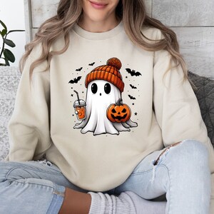Op de afbeelding: Beige sweatshirt met een cartoon spook met een oranje beanie, een pompoen en een drankje. Zwarte vleermuizen en sterren omringen het spook, wat een Halloween-thema cre&euml;ert.