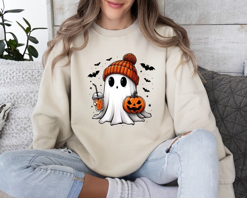 Op de afbeelding: Beige sweatshirt met een cartoon spook met een oranje beanie, een pompoen en een drankje. Zwarte vleermuizen en sterren omringen het spook, wat een Halloween-thema cre&euml;ert.