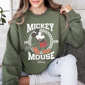 Op de afbeelding: Een olijfgroene sweatshirt met een Mickey Mouse-afbeelding. Het ontwerp bevat de woorden "MICKEY MOUSE" in een vintage lettertype, het jaar "1928" en het Disney-logo. Het stripfiguur staat in een geruite cirkel.