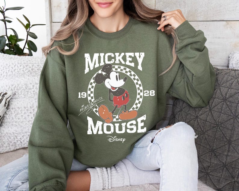 Op de afbeelding: Een olijfgroene sweatshirt met een Mickey Mouse-afbeelding. Het ontwerp bevat de woorden "MICKEY MOUSE" in een vintage lettertype, het jaar "1928" en het Disney-logo. Het stripfiguur staat in een geruite cirkel.