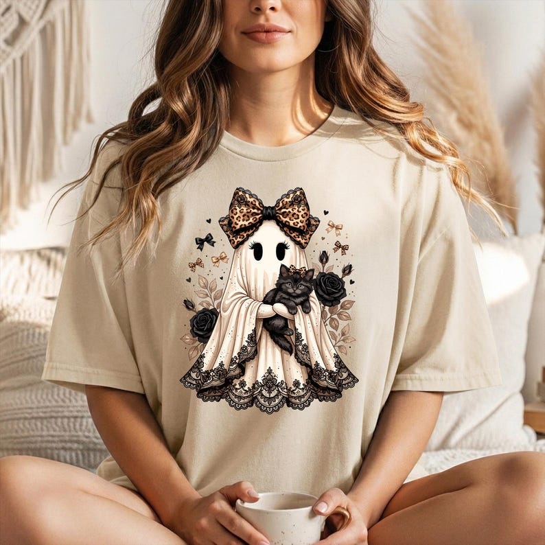 K&ouml;nnte beinhalten: Ein beiges T-Shirt mit kurzen &Auml;rmeln und einer Grafik eines Geistes, der eine gro&szlig;e Schleife mit Leopardenmuster tr&auml;gt und ein schwarzes K&auml;tzchen h&auml;lt. Der Geist ist mit schwarzer Spitzenborte am Saum verziert und von schwarzen Rosen umgeben.