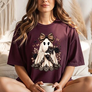K&ouml;nnte beinhalten: Ein dunkelweinrotes Kurzarm-T-Shirt mit einer Grafik eines wei&szlig;en Geistes, der eine Schleife mit Leopardenmuster tr&auml;gt und ein schwarzes K&auml;tzchen h&auml;lt. Der Geist ist mit schwarzer Spitzenborte am Saum verziert und von schwarzen Rosen und kleinen goldenen Schleifen umgeben.