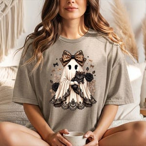 K&ouml;nnte beinhalten: Ein beiges T-Shirt mit kurzen &Auml;rmeln und einem Grafikdruck eines Geistes, der eine Schleife mit Leopardenmuster tr&auml;gt und ein schwarzes K&auml;tzchen h&auml;lt. Der Geist ist am Saum mit schwarzer Spitze verziert und von schwarzen Rosen umgeben.