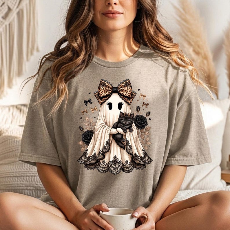 K&ouml;nnte beinhalten: Ein beiges T-Shirt mit kurzen &Auml;rmeln und einem Grafikdruck eines Geistes, der eine Schleife mit Leopardenmuster tr&auml;gt und ein schwarzes K&auml;tzchen h&auml;lt. Der Geist ist am Saum mit schwarzer Spitze verziert und von schwarzen Rosen umgeben.