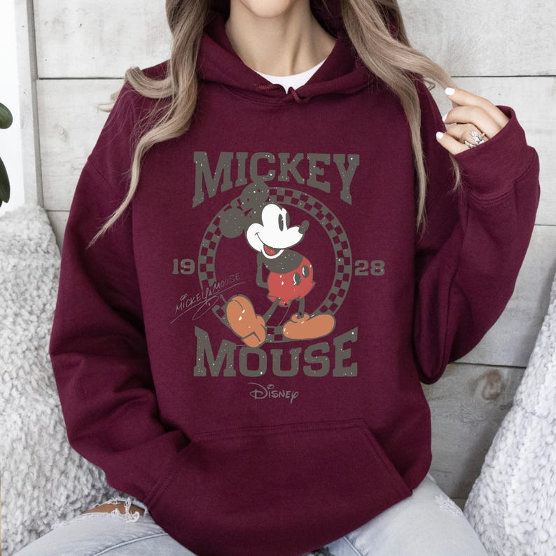 May include: Burgundy hoodie featuring a classic Mickey Mouse graphic. The design includes the words "MICKEY MOUSE" and "Disney" with the year "1928". The hoodie has a front pocket and a drawstring hood.