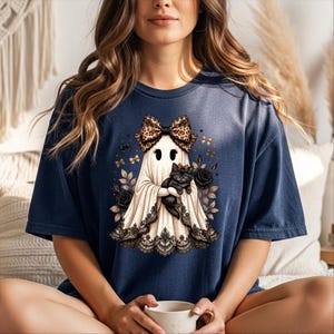 K&ouml;nnte beinhalten: Ein marineblaues T-Shirt mit einer Grafik eines wei&szlig;en Geistes, der eine Schleife mit Leopardenmuster tr&auml;gt und ein schwarzes K&auml;tzchen h&auml;lt. Der Geist ist am Saum mit schwarzer Spitze verziert und von schwarzen Rosen und kleinen goldenen Schmetterlingen umgeben.