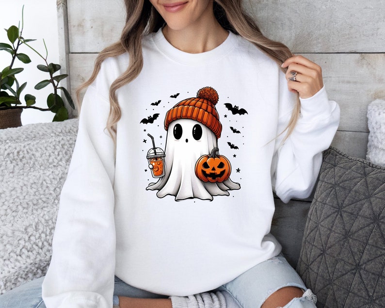 Op de afbeelding: Wit sweatshirt met een cartoongeest met een oranje gebreide muts. De geest houdt een pompoen en een drankje met een rietje vast. Zwarte vleermuizen en sterren omringen de geest en cre&euml;ren een Halloween-thema.