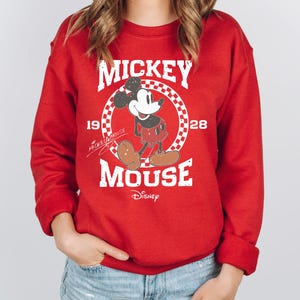 Op de afbeelding: Rode sweatshirt met een Mickey Mouse-afbeelding. Het ontwerp bevat de tekst "Mickey Mouse" in het wit, met het personage in een geruite cirkel. De sweatshirt heeft ook het jaartal "1928" en het Disney-logo.