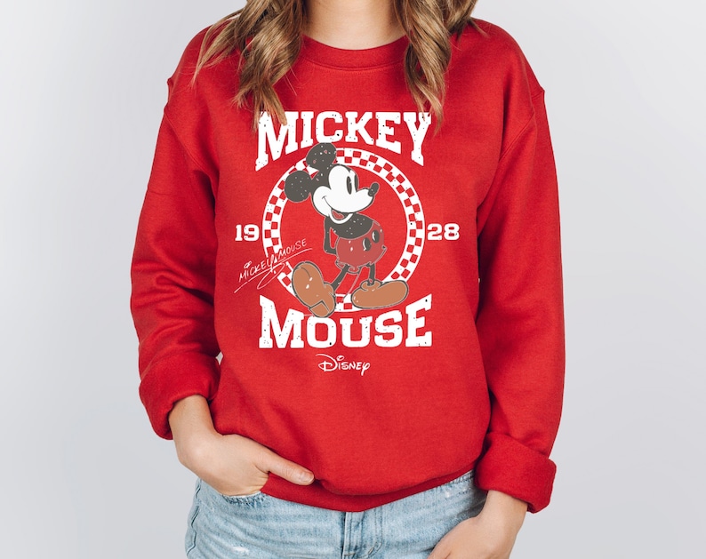 Op de afbeelding: Rode sweatshirt met een Mickey Mouse-afbeelding. Het ontwerp bevat de tekst "Mickey Mouse" in het wit, met het personage in een geruite cirkel. De sweatshirt heeft ook het jaartal "1928" en het Disney-logo.