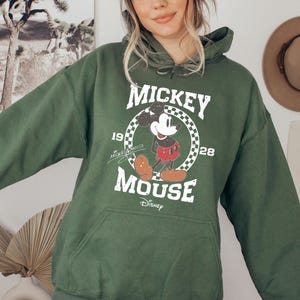 Op de afbeelding: Een groene hoodie met een Mickey Mouse-afbeelding en de tekst "Mickey Mouse 1928 Disney" gedrukt op de voorkant.