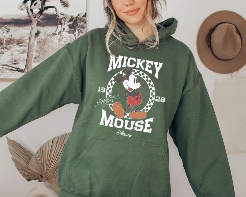 Op de afbeelding: Een groene hoodie met een Mickey Mouse-afbeelding en de tekst "Mickey Mouse 1928 Disney" gedrukt op de voorkant.