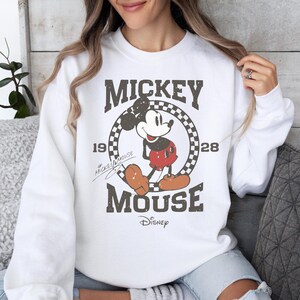 May include: White sweatshirt featuring a distressed graphic of Mickey Mouse. The design includes the text "Mickey Mouse 1928" in a vintage style, with the Disney logo at the bottom. The sweatshirt is a classic crew neck style.
