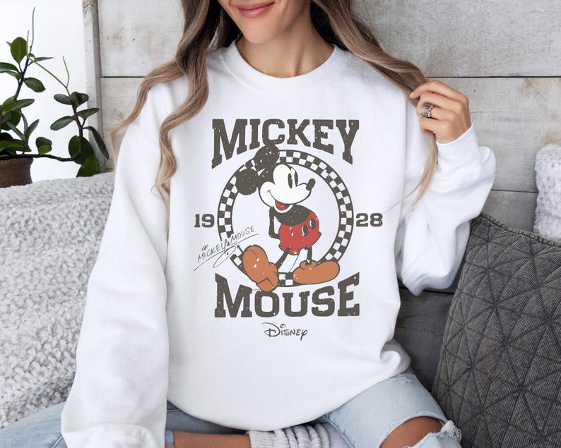 May include: White sweatshirt featuring a distressed graphic of Mickey Mouse. The design includes the text "Mickey Mouse 1928" in a vintage style, with the Disney logo at the bottom. The sweatshirt is a classic crew neck style.