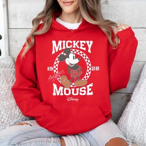 Op de afbeelding: Rood hoodie met een witte afbeelding van Mickey Mouse in een geruite cirkel. De afbeelding zegt "Mickey Mouse" met de jaren "1928" en "1928" aan weerszijden. De tekst "Disney" staat onder de afbeelding.