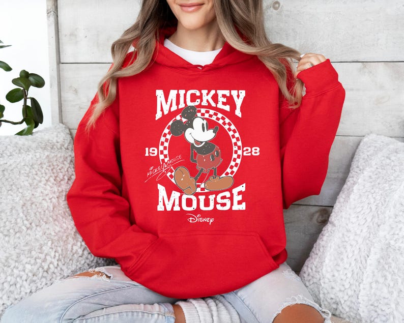 Op de afbeelding: Rood hoodie met een witte afbeelding van Mickey Mouse in een geruite cirkel. De afbeelding zegt "Mickey Mouse" met de jaren "1928" en "1928" aan weerszijden. De tekst "Disney" staat onder de afbeelding.