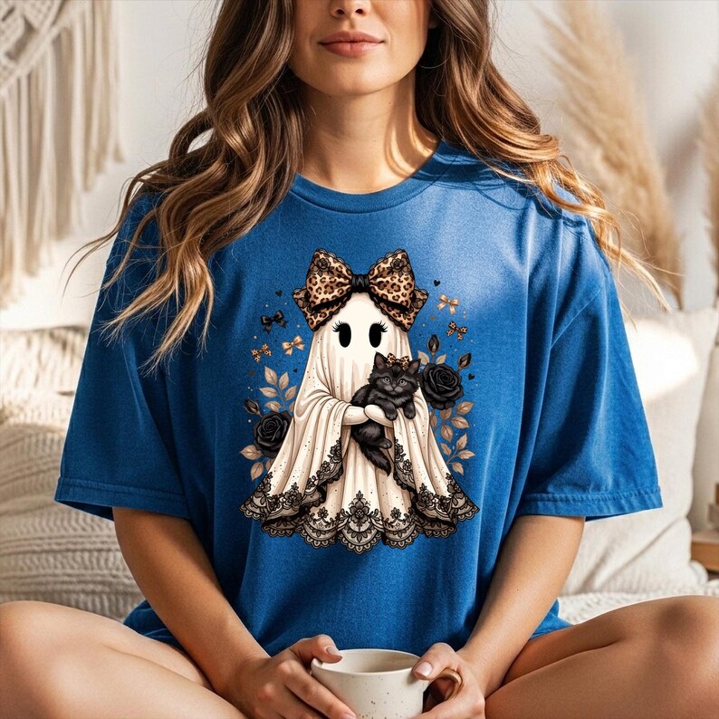 K&ouml;nnte beinhalten: Ein blaues T-Shirt mit einer Grafik eines wei&szlig;en Geistes, der eine Schleife mit Leopardenmuster tr&auml;gt und ein schwarzes K&auml;tzchen h&auml;lt. Der Geist ist von schwarzen Rosen und goldenen Bl&auml;ttern umgeben, mit einem schwarzen Spitzenbesatz am Saum.