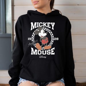Op de afbeelding: Zwart hoodie met een vintage Mickey Mouse-print. De print toont Mickey Mouse in een rood-witte outfit, staand in een cirkel met de tekst "Mickey Mouse" en "1928" eromheen geschreven. De tekst "Disney" staat onder de cirkel.