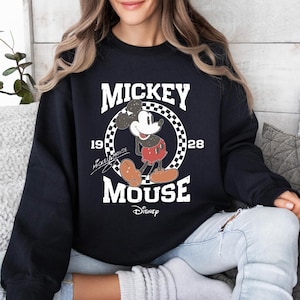 Op de afbeelding: Zwart sweatshirt met een vintage Mickey Mouse-ontwerp. Het ontwerp bevat de woorden "MICKEY MOUSE" en "1928" in het wit, samen met een afbeelding van Mickey Mouse. Het Disney-logo staat onderaan.