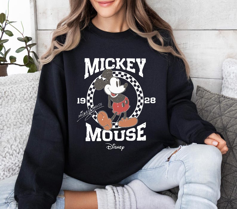 Op de afbeelding: Zwart sweatshirt met een vintage Mickey Mouse-ontwerp. Het ontwerp bevat de woorden "MICKEY MOUSE" en "1928" in het wit, samen met een afbeelding van Mickey Mouse. Het Disney-logo staat onderaan.