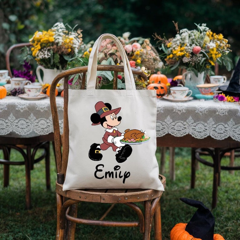 K&ouml;nnte beinhalten: Eine wei&szlig;e Canvas-Tragetasche mit einem Cartoon-Bild von Mickey Mouse, der als Pilger verkleidet ist und einen Teller mit Essen tr&auml;gt. Die Tasche tr&auml;gt den Namen "Emily" auf der Unterseite.