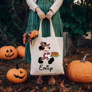 K&ouml;nnte beinhalten: Eine wei&szlig;e Canvas-Tragetasche mit einem Thanksgiving-Motiv, auf dem Mickey Mouse als Pilger dargestellt ist, der einen Teller mit Essen tr&auml;gt. Die Tasche tr&auml;gt den Namen "Emily" auf der Unterseite.