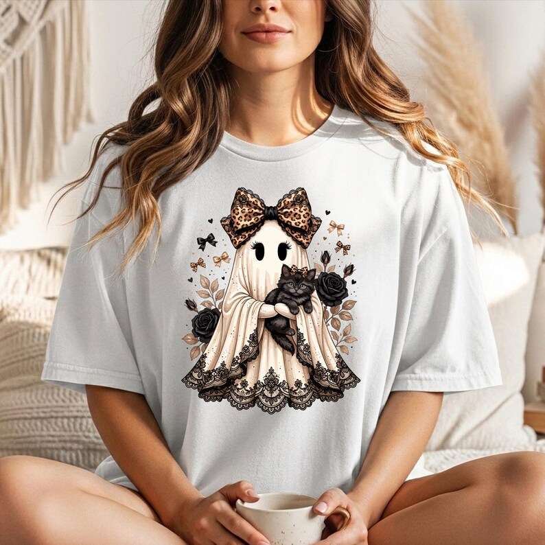 K&ouml;nnte beinhalten: Ein wei&szlig;es T-Shirt mit einer Grafik eines Geistes, der eine gro&szlig;e Schleife mit Leopardenmuster tr&auml;gt und ein schwarzes K&auml;tzchen h&auml;lt. Der Geist ist am Saum mit schwarzer Spitze verziert und von schwarzen Rosen, kleinen Schleifen und Bl&auml;ttern umgeben.