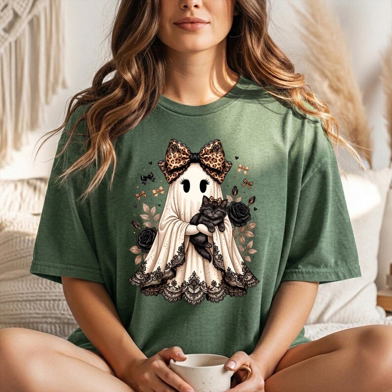 K&ouml;nnte beinhalten: Ein olivgr&uuml;nes T-Shirt mit einer Grafik eines wei&szlig;en Geistes, der eine Schleife mit Leopardenmuster tr&auml;gt und ein schwarzes K&auml;tzchen h&auml;lt. Der Geist ist am Saum mit schwarzer Spitze verziert und von schwarzen Rosen, Bl&auml;ttern und kleinen Schleifen umgeben.