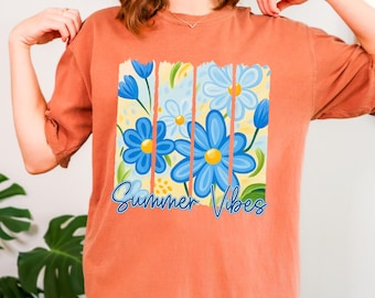 May include: Orange t-shirt featuring a floral design with blue flowers and the text "Summer Vibes" in a script font. The flowers have yellow centers and are set against a yellow and light blue background.