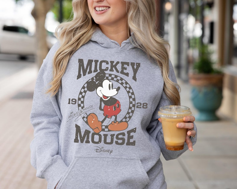 Op de afbeelding: Een grijze hoodie met een vintage Mickey Mouse-print. De print toont Mickey Mouse in een geruite cirkel met de tekst "Mickey Mouse" en "1928" boven en onder de cirkel. De tekst "Disney" staat onder de cirkel.