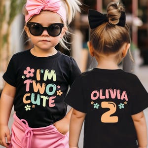 Puede incluir: Dos ni&ntilde;as con camisetas negras con texto de colores. Una camiseta dice "I'm Two Cute" y la otra dice "Olivia 2" con flores.