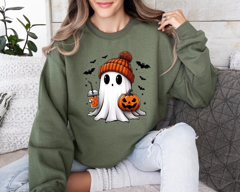 Op de afbeelding: Olijfgroene sweatshirt met een cartoon spookontwerp. Het spook draagt een oranje muts en houdt een pompoen en een drankje vast. Zwarte vleermuizen en sterren zijn verspreid rond het spook. Het spook heeft grote zwarte ogen en een vriendelijke uitdrukking.