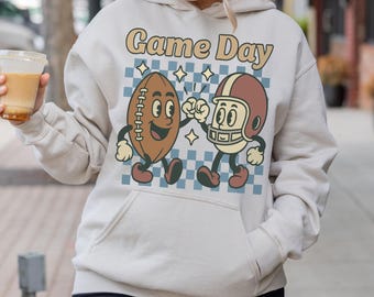 May include: A cream-colored hoodie with a front pocket and the words "Game Day" in a retro font. The hoodie features a cartoon illustration of a football and a football helmet giving a high-five, set against a blue and white checkered background.