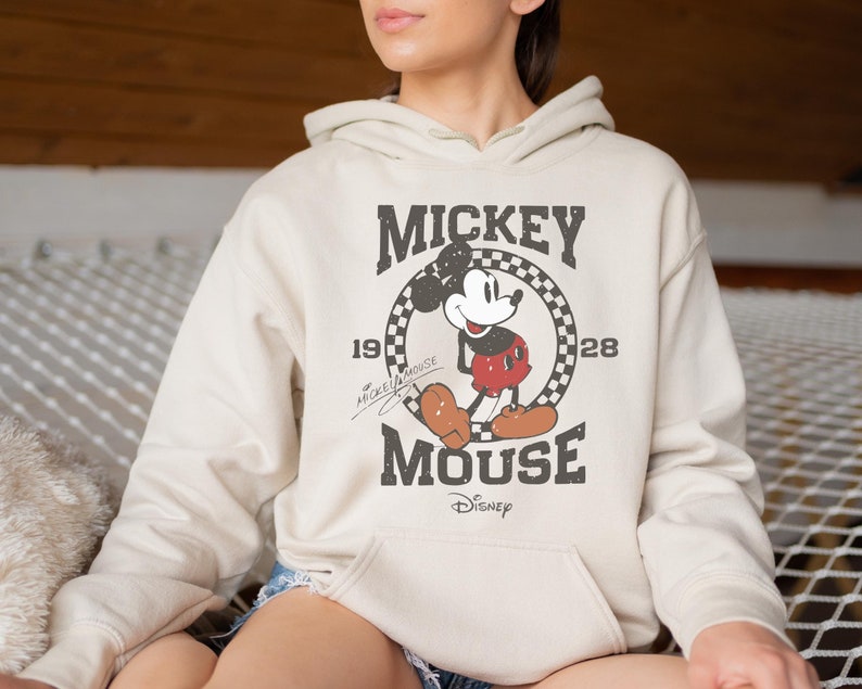 Op de afbeelding: Een cr&egrave;mekleurige hoodie met een vintage Mickey Mouse-afbeelding. De afbeelding toont Mickey Mouse in een rood-zwarte outfit, met de tekst "Mickey Mouse" en "1928" in een zwart-wit geblokte cirkel.