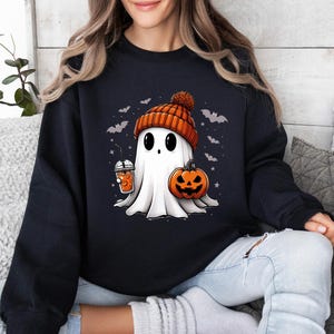 Op de afbeelding: Zwart sweatshirt met een cartoongeest met een oranje muts, een pompoen en een drankje. De geest heeft grote zwarte ogen en is omringd door vleermuizen en sterren, perfect voor Halloween.