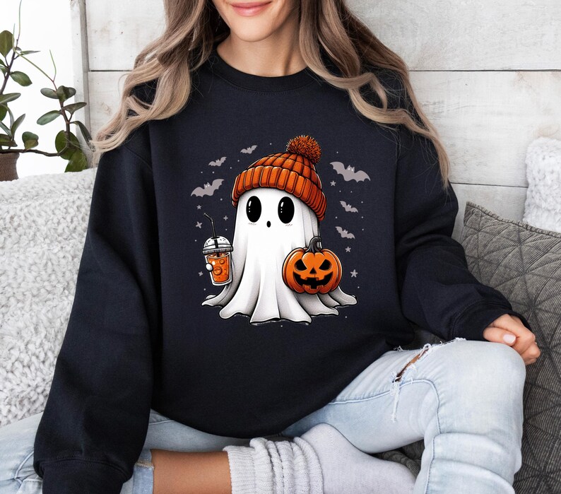 Op de afbeelding: Zwart sweatshirt met een cartoongeest met een oranje muts, een pompoen en een drankje. De geest heeft grote zwarte ogen en is omringd door vleermuizen en sterren, perfect voor Halloween.