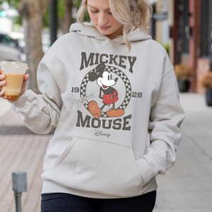 May include: A light beige hoodie featuring a vintage Mickey Mouse graphic. The graphic includes the words "Mickey Mouse" and "Disney", along with the years "19" and "28". The hoodie has a front pocket and a drawstring hood.