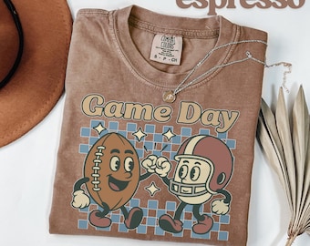 May include: Espresso-colored t-shirt with a retro "Game Day" graphic featuring a cartoon football and a football helmet character. The design includes a blue and white checkered pattern and star accents. A silver necklace is draped across the shirt.