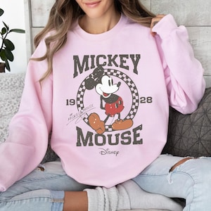 Op de afbeelding: Lichtroze sweatshirt met een verweerde Mickey Mouse-afbeelding. Het ontwerp bevat de tekst "Mickey Mouse" en het jaar "1928" in een geruite cirkel. Het Disney-logo staat onderaan.