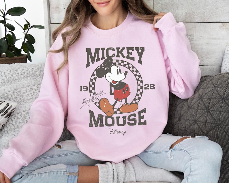 Op de afbeelding: Lichtroze sweatshirt met een verweerde Mickey Mouse-afbeelding. Het ontwerp bevat de tekst "Mickey Mouse" en het jaar "1928" in een geruite cirkel. Het Disney-logo staat onderaan.