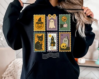 May include: Black hooded sweatshirt featuring a Halloween-themed stamp design. The stamps depict a jack-o'-lantern, ghost, lollipop, black cat, haunted house, and skull with a tombstone. The design is in orange, purple, green, and yellow.