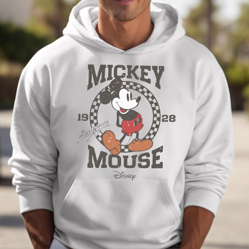 May include: White hooded sweatshirt featuring a distressed graphic of Mickey Mouse. The design includes the text "Mickey Mouse" above and below a circular checkered pattern with the year "1928". The Disney logo is at the bottom.