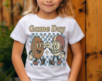 May include: White t-shirt featuring a cartoon football and helmet with the text "Game Day". The design includes a brown football with a smiling face and a helmet with a maroon helmet. The background has a blue and white checkered pattern.