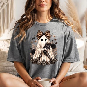 K&ouml;nnte beinhalten: Ein graues T-Shirt mit einem Grafikdruck eines Geistes, der eine Schleife mit Leopardenmuster tr&auml;gt und ein schwarzes K&auml;tzchen h&auml;lt. Der Geist ist am Saum mit schwarzer Spitze verziert und von schwarzen Rosen sowie kleinen goldenen Schleifen umgeben.