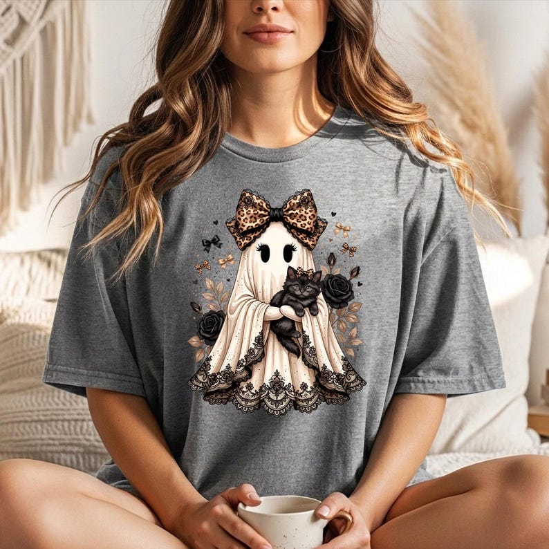 K&ouml;nnte beinhalten: Ein graues T-Shirt mit einem Grafikdruck eines Geistes, der eine Schleife mit Leopardenmuster tr&auml;gt und ein schwarzes K&auml;tzchen h&auml;lt. Der Geist ist am Saum mit schwarzer Spitze verziert und von schwarzen Rosen sowie kleinen goldenen Schleifen umgeben.