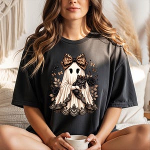 K&ouml;nnte beinhalten: Ein anthrazitfarbenes T-Shirt mit einer Grafik eines wei&szlig;en Geistes, der eine Schleife mit Leopardenmuster tr&auml;gt und eine schwarze Katze h&auml;lt. Der Geist ist am Saum mit schwarzer Spitze verziert und von schwarzen Rosen und kleinen goldenen Schleifen umgeben.