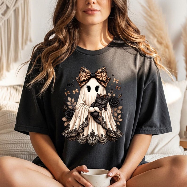 K&ouml;nnte beinhalten: Ein anthrazitfarbenes T-Shirt mit einer Grafik eines wei&szlig;en Geistes, der eine Schleife mit Leopardenmuster tr&auml;gt und eine schwarze Katze h&auml;lt. Der Geist ist am Saum mit schwarzer Spitze verziert und von schwarzen Rosen und kleinen goldenen Schleifen umgeben.