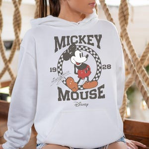 Op de afbeelding: Witte hoodie met een vintage Mickey Mouse-afbeelding. De afbeelding toont Mickey Mouse in een rood-witte outfit met de tekst "Mickey Mouse" en "1928" in een zwart-wit geblokte cirkel.